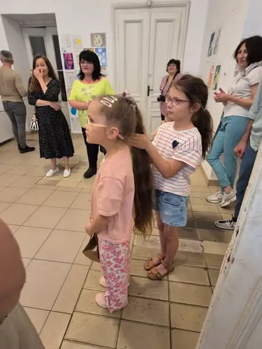 Две момичета стоят в коридор на изложбата, зад тях възрастни разглеждат рисунки.