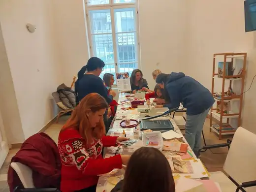 Хора работят край дълга маса в светла зала с високи прозорци.