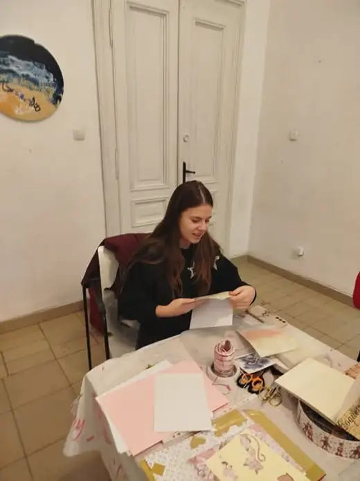 Момиче седи на маса с листове розова и жълта хартия и изработва картичка.