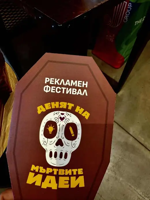 Ръка държи картонена табелка във формата на ковчег с череп и надпис „Денят на мъртвите идеи“.