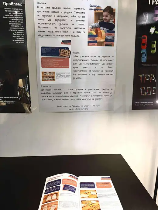 Плакат „Дигитално списание Светът на децата“ на стена, под него на маса лежи отворено списание „Тайнствените торбички“.