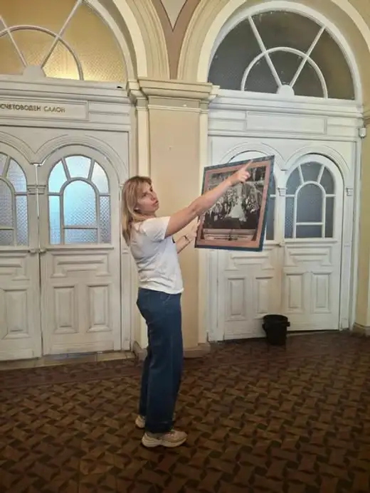 Жена в бяла тениска показва картина в рамка на група ученици.