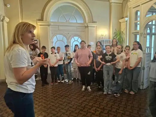 Ученици стоят в кръг и слушат жена в бяла тениска в зала с арковидни прозорци.