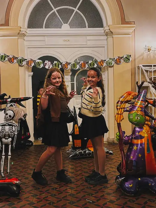 Две момичета с нарисуван грим позират в зала с гирлянда „Happy Halloween“ и балон-вещица.