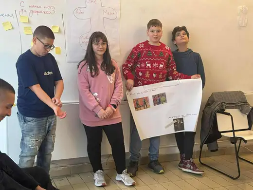 Четирима ученици стоят пред стена с лепящи листчета и държат голям лист с бележки.