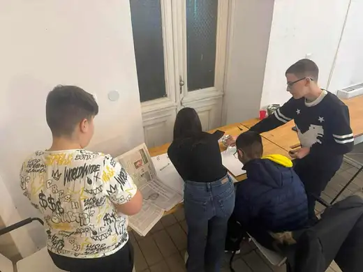 Момчета и момиче се навеждат над лист на маса до прозорец.