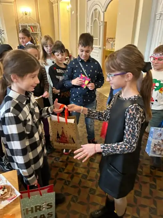 Момиче подава коледна подаръчна торбичка на съученичка, децата наоколо гледат.