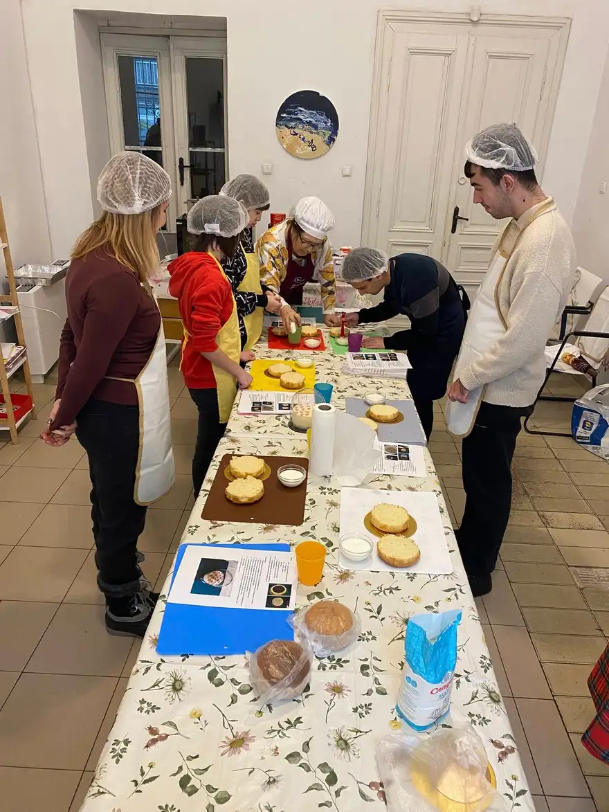 Няколко души със шапки за коса стоят край дълга маса с цветна мушама, върху която има форми, купички и печени блатове.