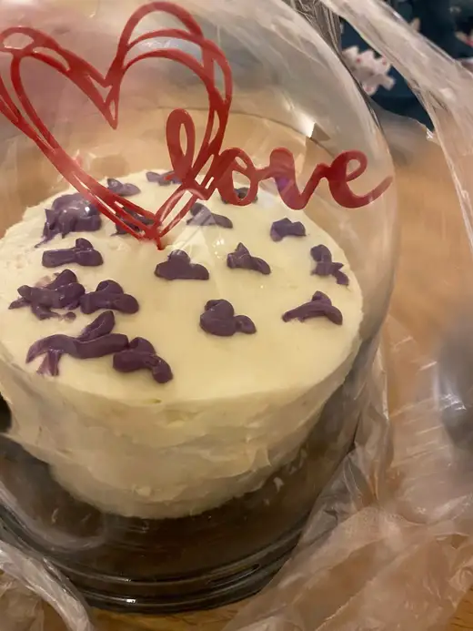 Торта с бял крем, лилави декорации и червен надпис „love“ под прозрачен капак.