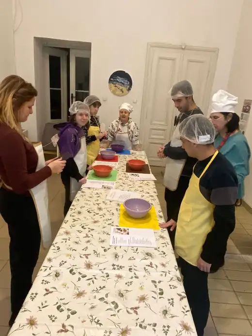 Група хора със шапки за коса и престилки край дълга маса, подредена с рецепти и съдове.