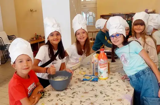 Група деца с бели готварски шапки се усмихват край маса с купа с тесто и бутилка мляко.