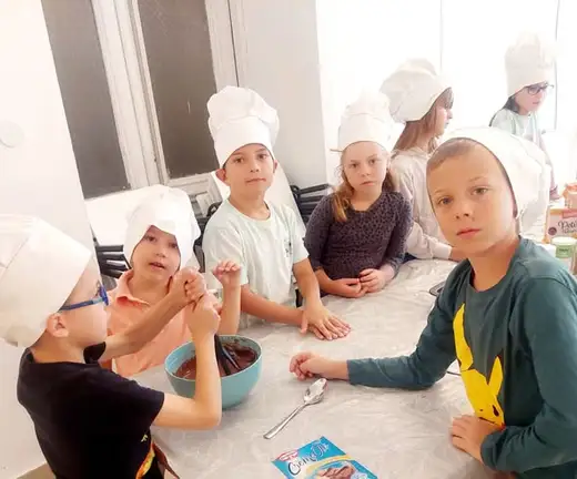 Деца с бели готварски шапки чакат край синя купа с разтопен шоколад и лъжица.