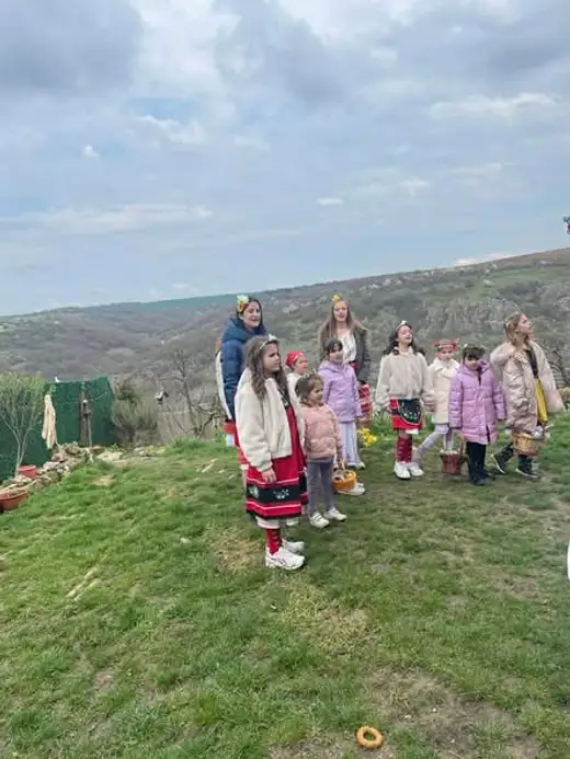 Момичета в носии и зимни якета пеят на тревиста поляна над скалист каньон.