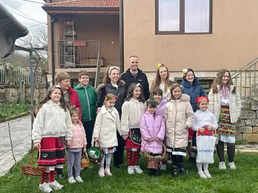 Групова снимка на децата с венчета и кошници заедно с придружителите им пред селска къща.
