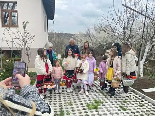 Възрастен снима с телефон групата лазарки с кошници в двор с цъфнали сливи.