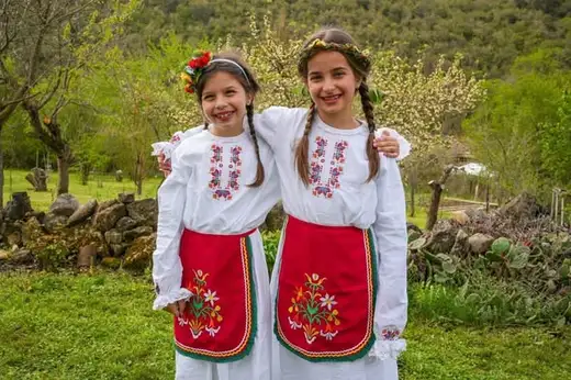 Две момичета в бели ризи с бродерия и червени престилки, прегърнати на тревиста поляна.