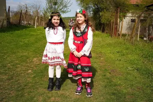 Две момичета с венчета и народни носии стоят хванати за ръце на тревиста поляна.