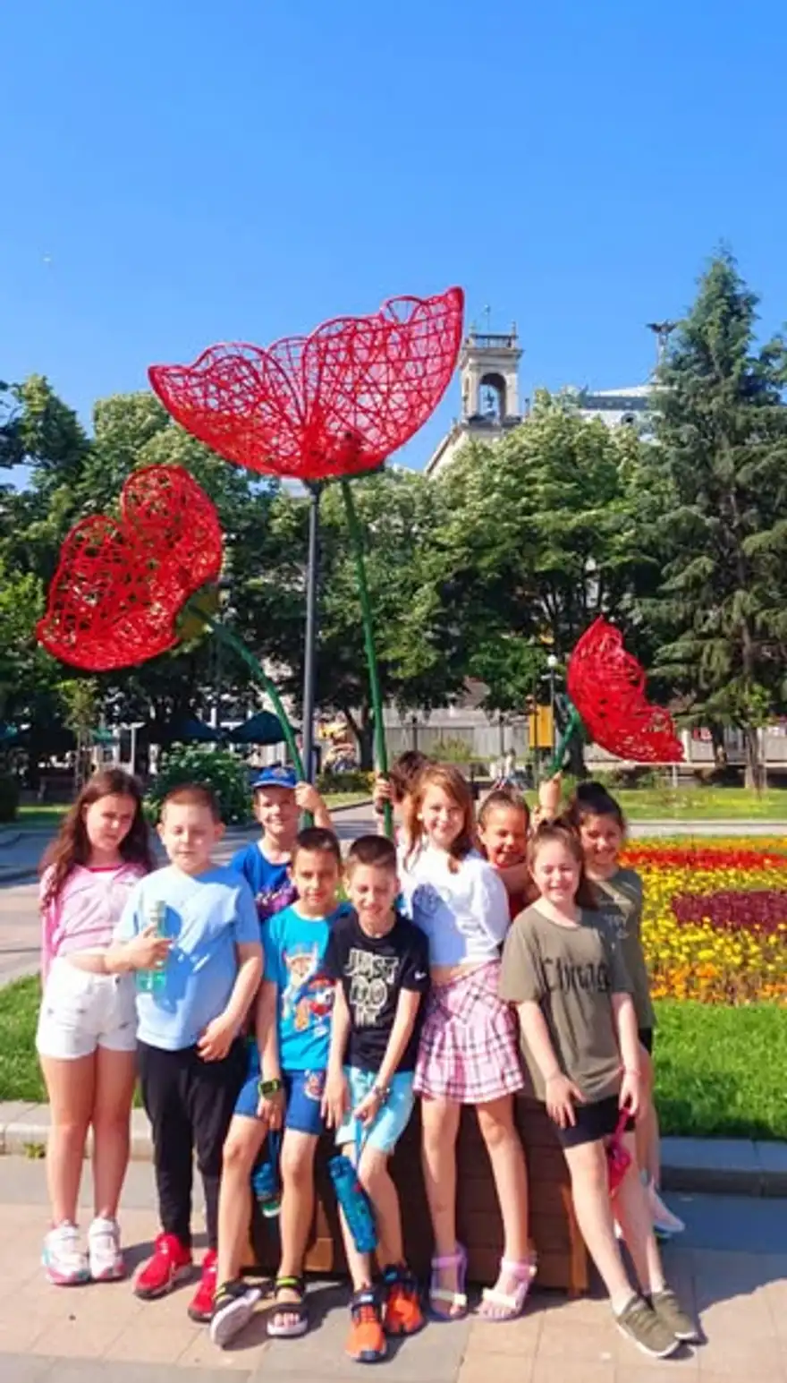 Група деца позира пред цветна леха с големи скулптури на червени макове.