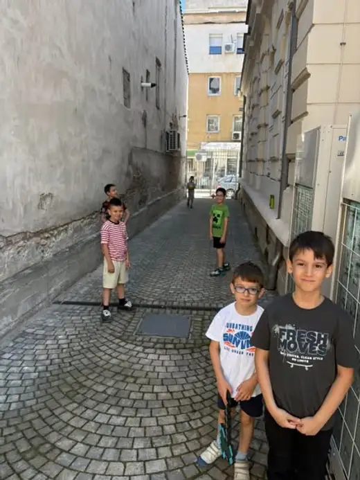 Две момчета стоят на павирана уличка между сгради, зад тях още деца.