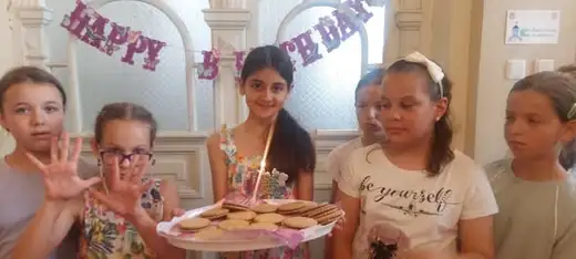 Момиче държи чиния с курабии и запалена свещичка, около него деца, а зад тях надпис „Happy Birthday“.