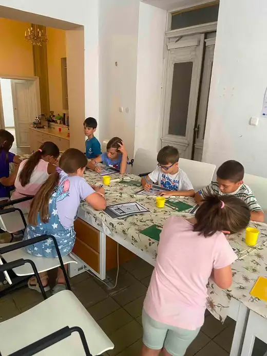 Деца работят по зелени подложки с глина на маса с флорална покривка.