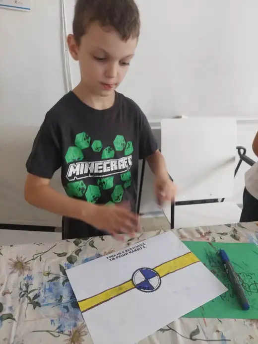Момче в тениска с надпис Minecraft оцветява знаме с маркер на маса.