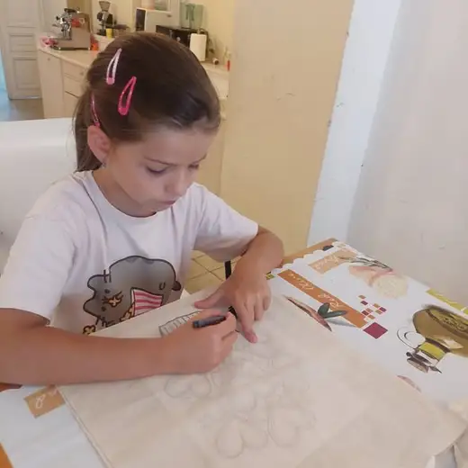 Момиче с щипка за коса рисува с молив върху лист на масата.