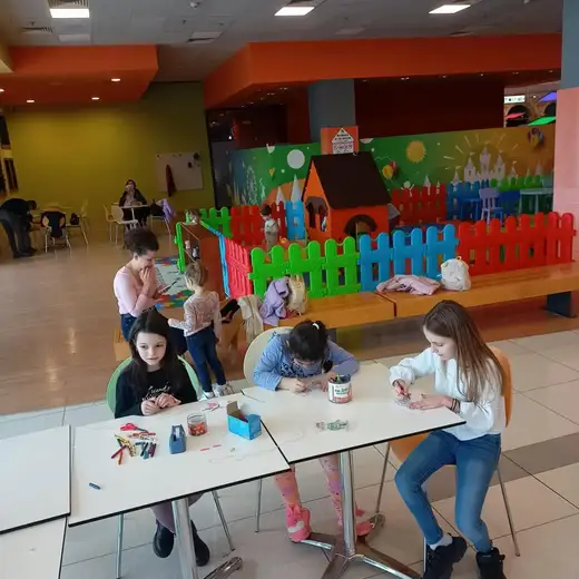 Група деца работят на маси в мола, зад тях цветна детска площадка с оградка.