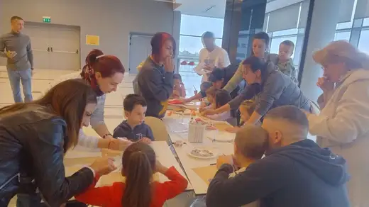 Деца и възрастни са се наведени над маси в мола и работят по есенни картини с маркери и стикери.