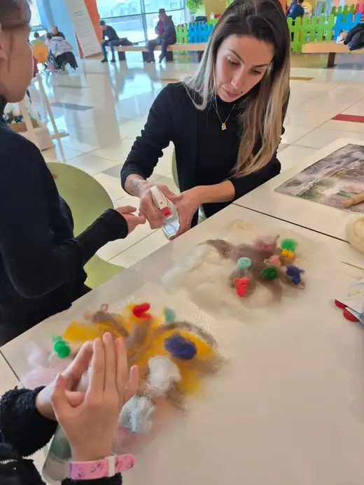 Жена пръска вода върху разпръснати цветни вълнени кичури върху бяла маса.