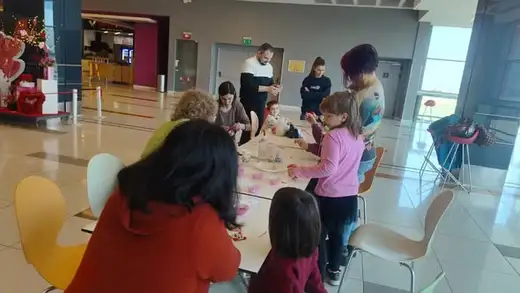 Възрастни и деца работят на обща маса в мола, на заден план има червени декорации.