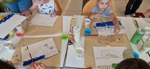 Изглед отгоре към маси с картони, чашки с вода, четки и започнати рисунки в синьо.