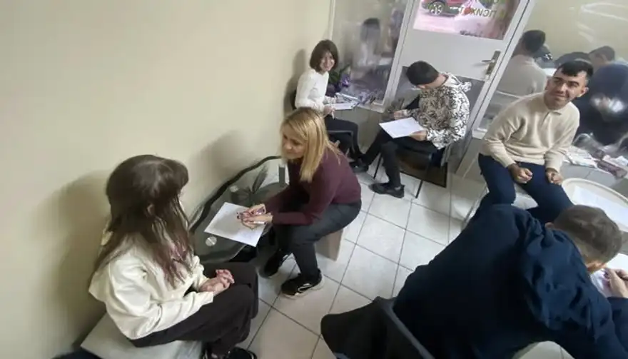Млади хора седят в кръг на столове в малка стая и пишат по листове, а жена с руса коса им говори.