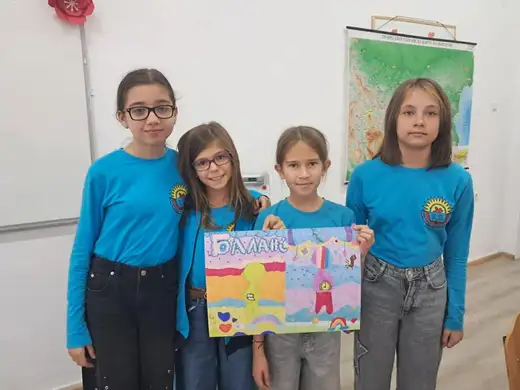 Четири ученички в тюркоазени блузи държат цветен плакат пред карта на стената.