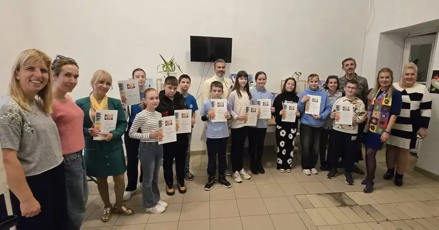 Група деца и възрастни в светла зала се снимат, като децата държат грамоти; на стената зад тях има рисунки.