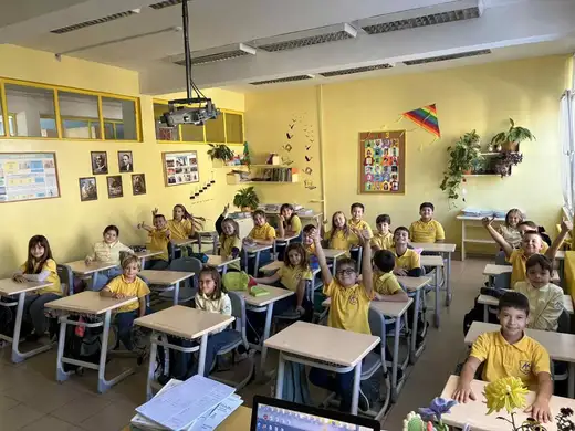 Ученици в жълти блузи седят на чиновете в класна стая, някои вдигат ръце.