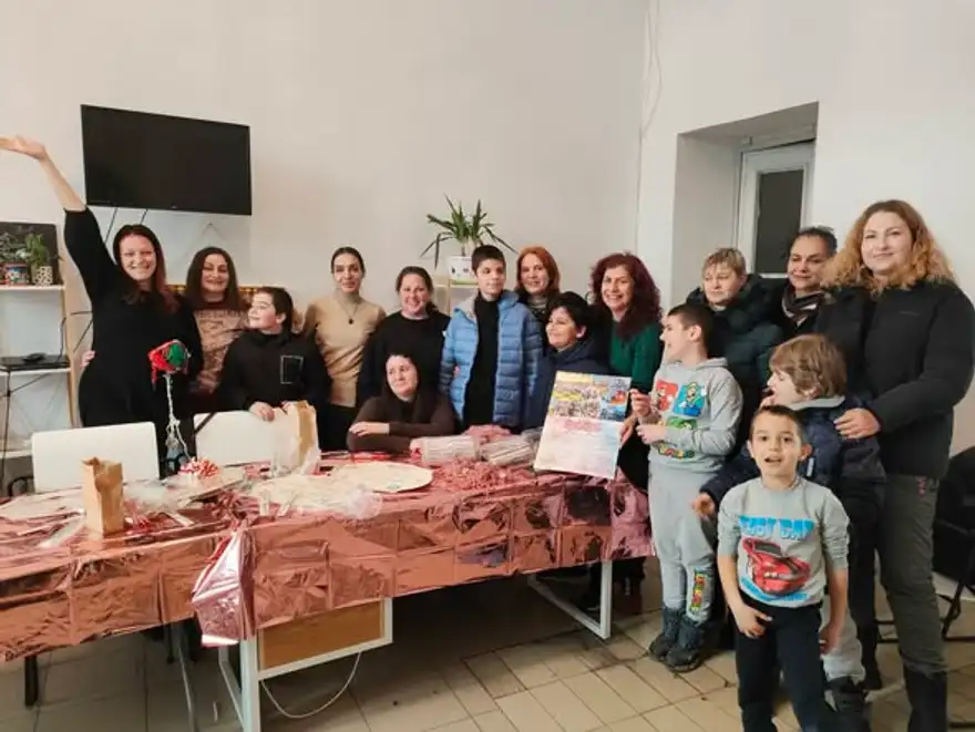 Група жени и деца стоят до маса, отрупана с ръчно изработени мартеници в червено и бяло.