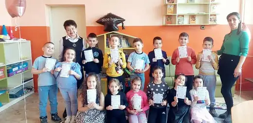 Клас деца с учителка позират в класната стая, повечето държат бели листчета.