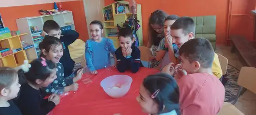 Деца седят около маса с червена покривка.