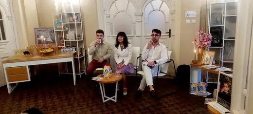Трима души седят на масичка в светла стая с рафтове с книги.