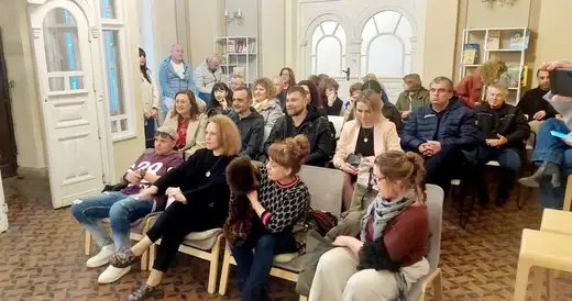 Зрители седят на столове в светлата зала.