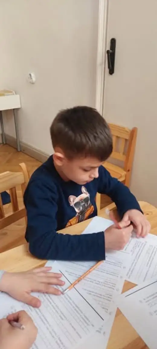 Момче попълва работен лист с молив, седнало край ниска дървена маса.