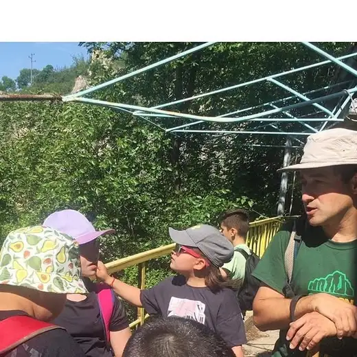 Деца с шапки и мъж в зелена тениска стоят под метална навесна конструкция, зад тях скала и жълт парапет.