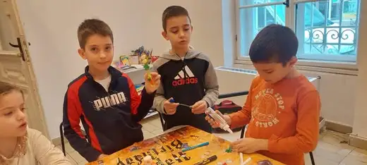 Три момчета работят с цветни материали на маса.