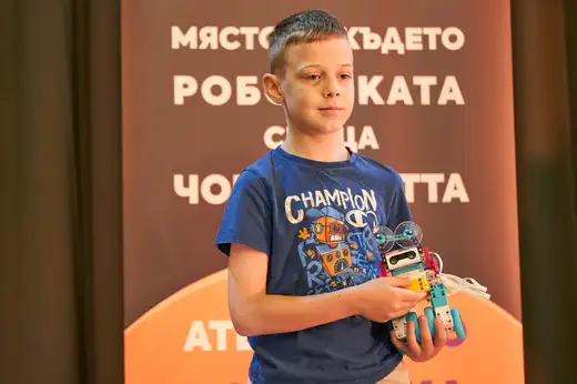 Момче държи сглобен робот пред оранжево пано с надпис „Мястото, където роботиката среща човека“.