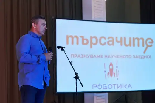 Мъж говори до екран с надпис „Търсачите“ и „Празнуваме наученото заедно“.