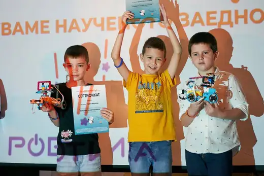 Три момчета вдигат робот и сертификат пред стена с надпис „Роботика“.