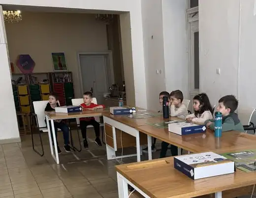 Просторна зала с дълга маса, около която седят деца с кутии от конструктор.