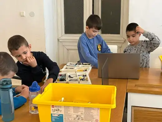 Три деца работят с елементи от конструктор около жълта кутия и отворен лаптоп.