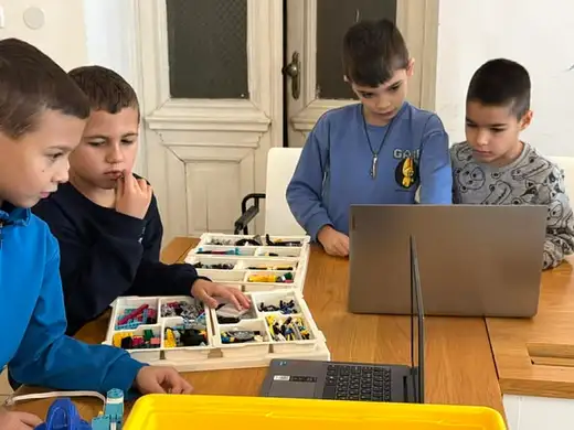 Момче въвежда нещо на лаптоп, а до него други две деца сглобяват от кутия с детайли.